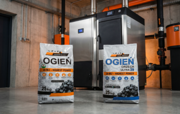 Dwa 25-kilogramowe worki węgla marki OGIEŃ stojące na betonowej podłodze w nowoczesnej kotłowni. W tle widoczny jest zaawansowany technologicznie piec c.o. ze stali nierdzewnej z panelami sterowania. Lewy worek ma pomarańczowe oznaczenia, prawy (Orzech Ultra 29) niebieskie. Oba opakowania informują o wysokiej kaloryczności 29 MJ – Highest Power.